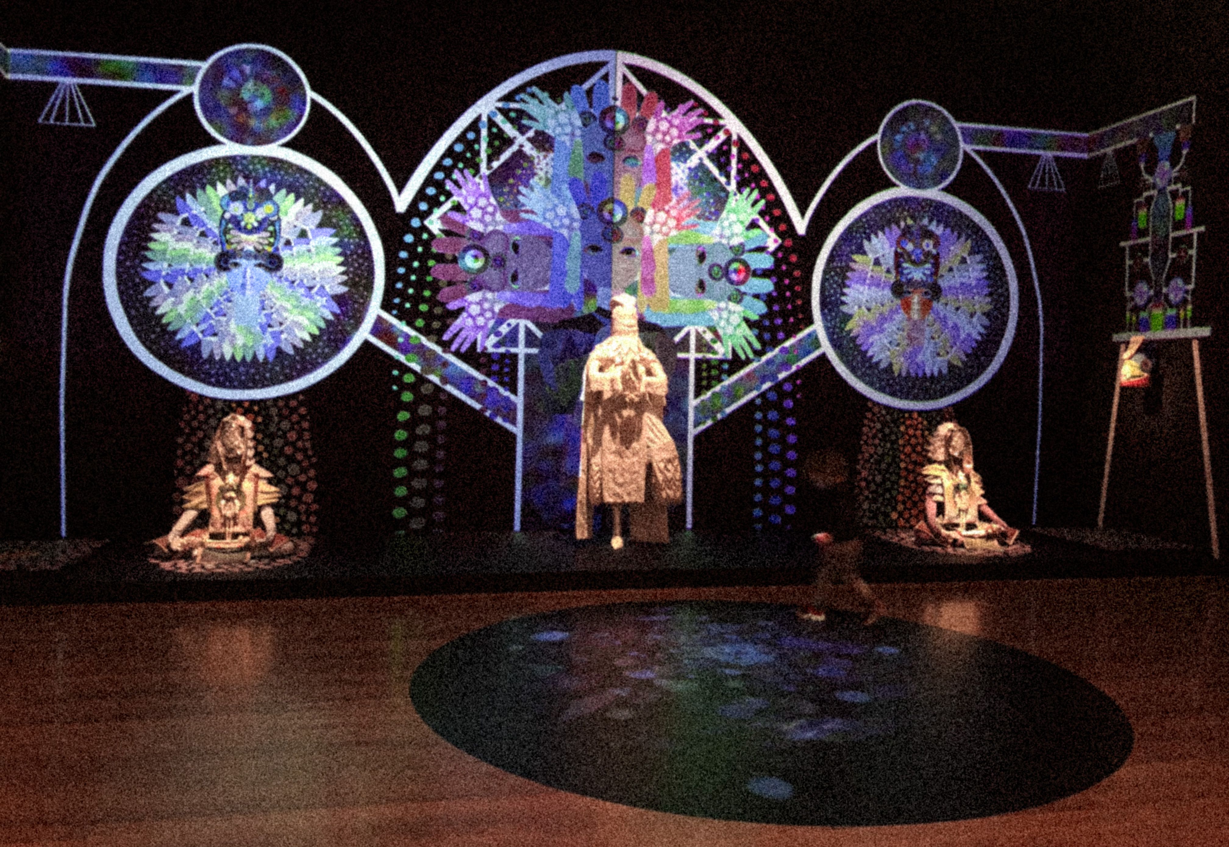 An art installation with a colorful geometric designs on the walls within what appears to be a series of tubes and containers of some kind. Two sculptures of people sit on the ground in meditative poses while a third statue stands on one leg with hands together. A child is walking on top of projected colorful shapes on the ground.